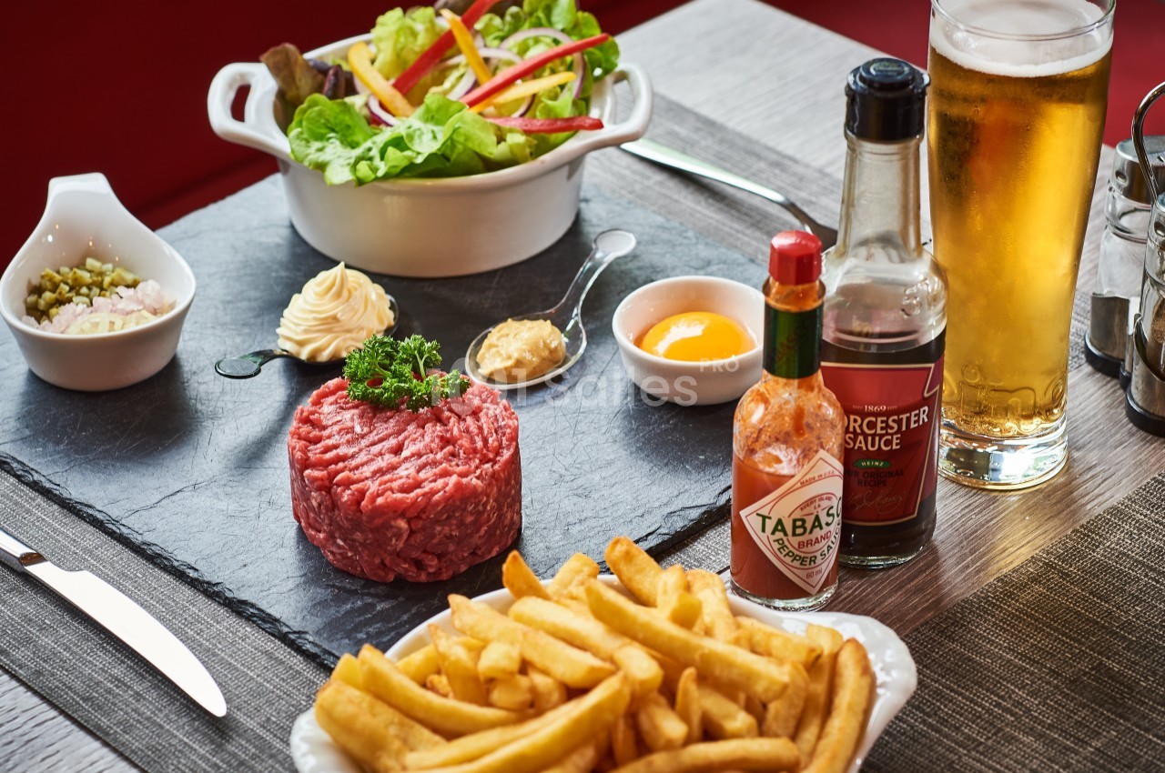 Assiette de tartare de bœuf cru avec frites, salade, condiments variés et verre de bière sur une table en bois.