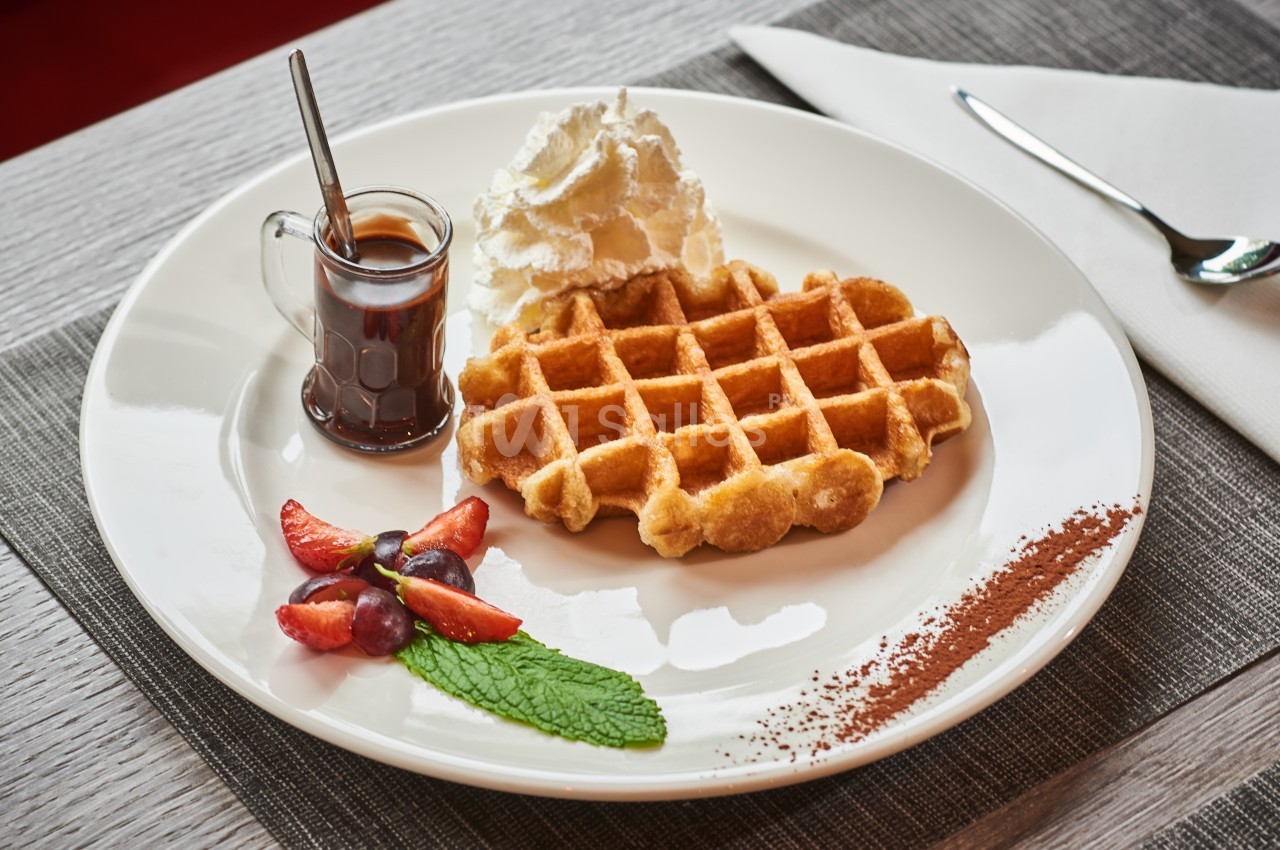 Assiette avec une gaufre, chantilly, sirop dans une cruche, fraises, raisins, feuille de menthe et poudre de cacao.