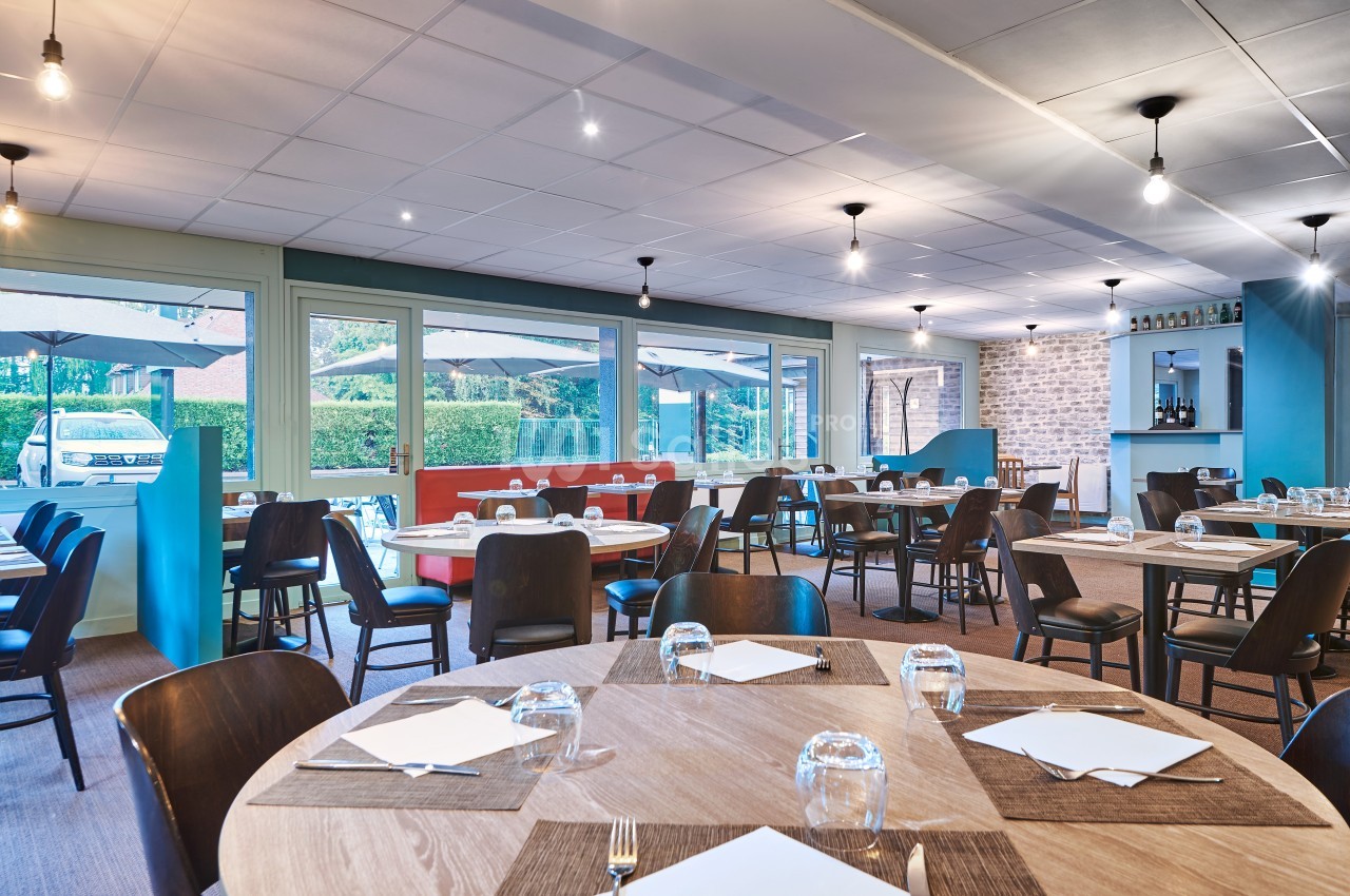 Salle de restaurant lumineuse avec tables dressées, grandes baies vitrées et vue sur une terrasse extérieure.