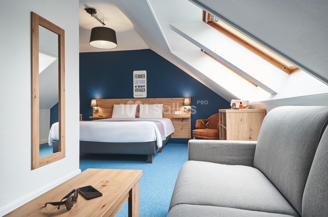 Chambre mansardée avec lit double, canapé gris, table basse en bois et lumière naturelle provenant de fenêtres de toit.