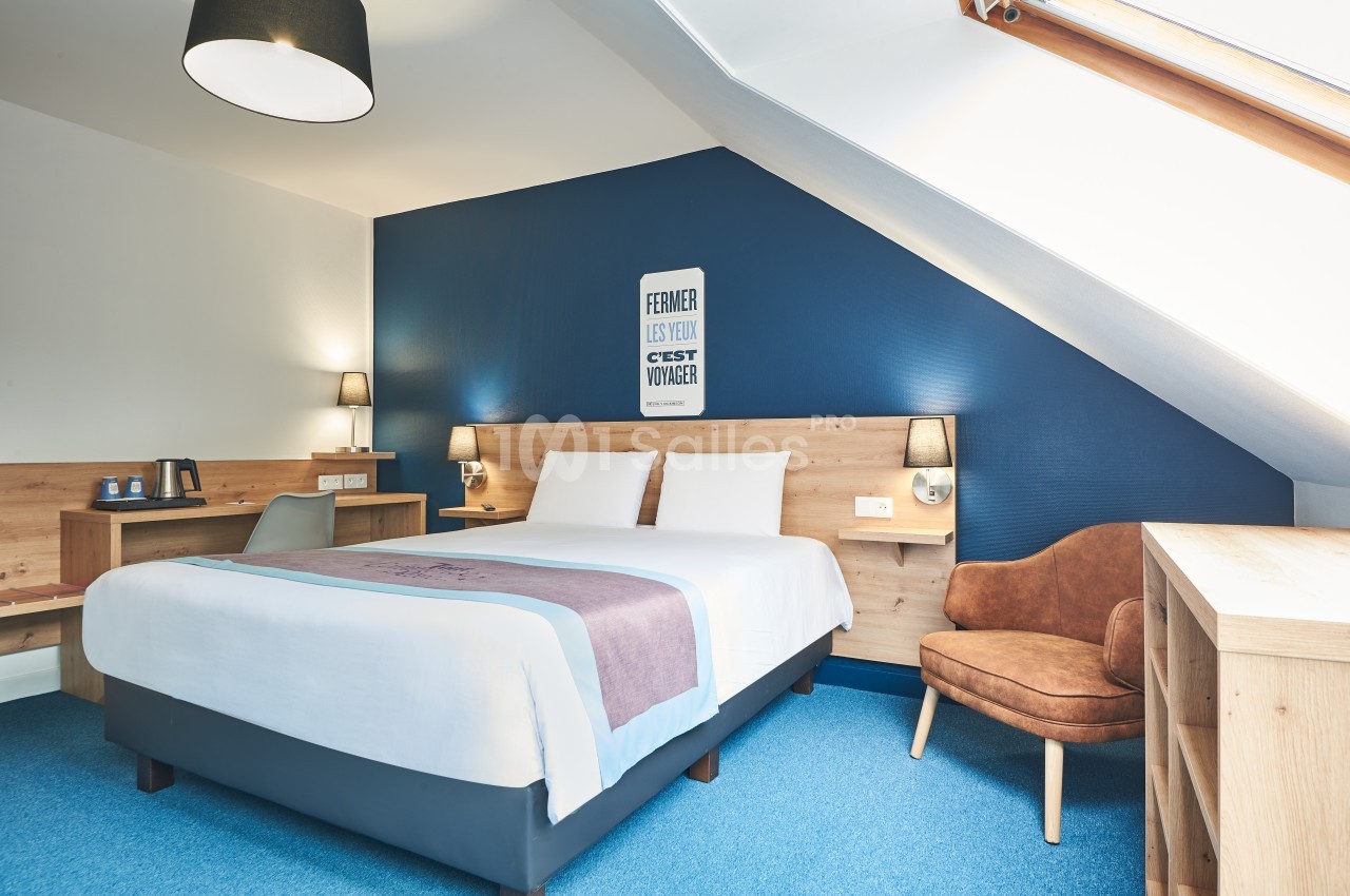 Chambre lumineuse avec lit double, mur bleu, bureau en bois, fauteuil et fenêtre de toit.