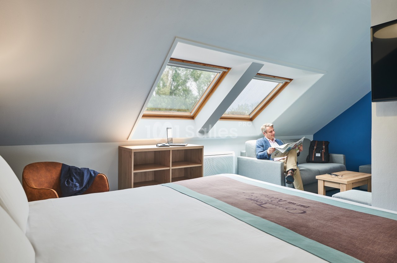 Homme assis dans un fauteuil sous des fenêtres de toit, lisant un journal dans une chambre lumineuse avec un lit au premier…