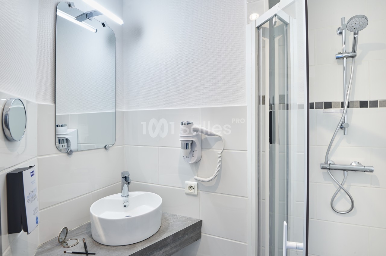 Salle de bain moderne avec lavabo, miroir éclairé, sèche-cheveux mural et cabine de douche avec pommeau réglable.