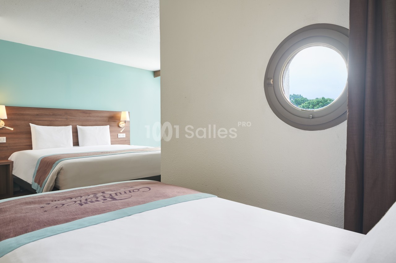 Deux lits dans une chambre d'hôtel lumineuse avec un hublot offrant une vue sur l'extérieur verdoyant.