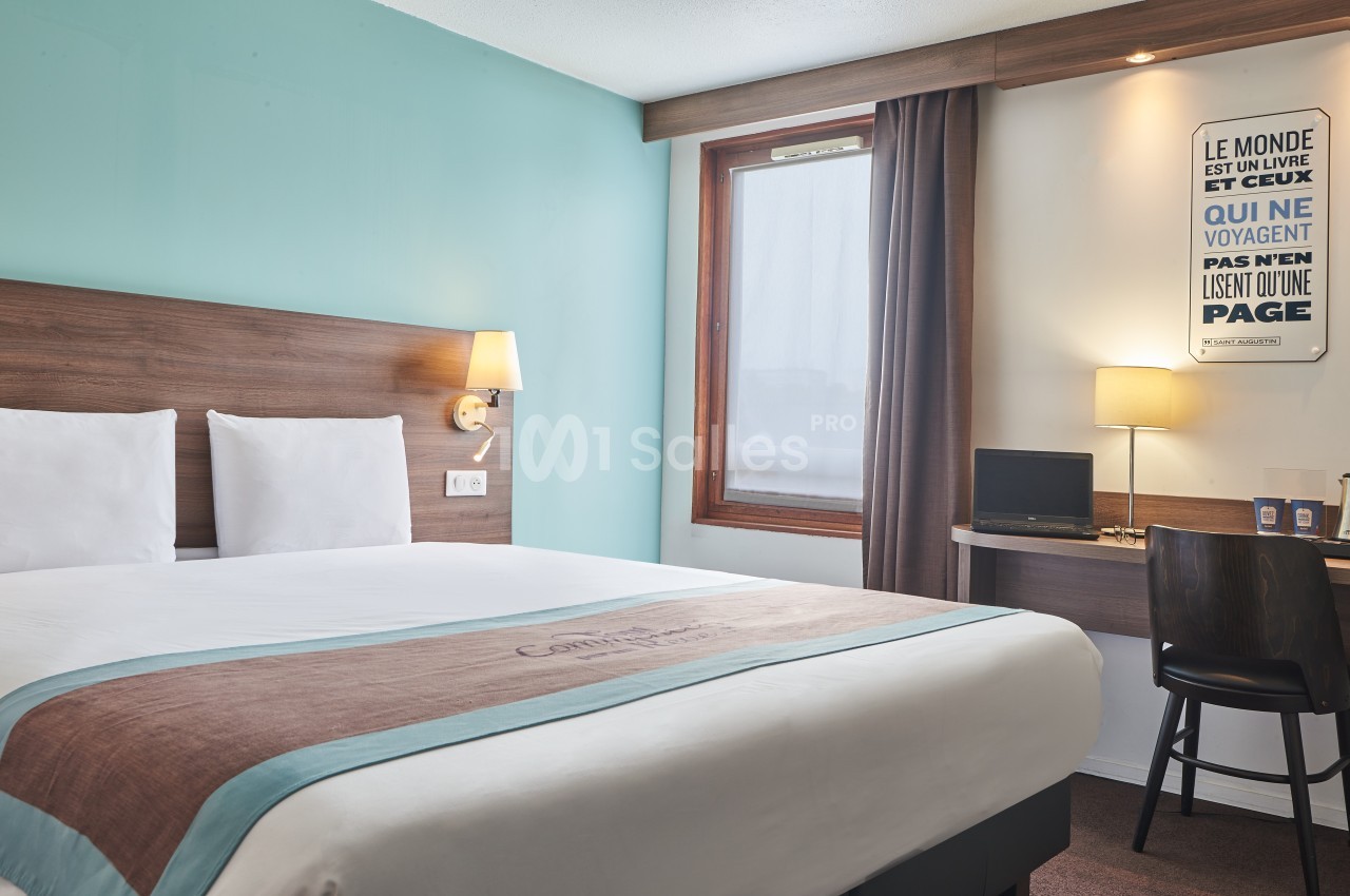 Chambre d'hôtel avec lit double, bureau équipé, lampe allumée et fenêtre avec rideaux marron.