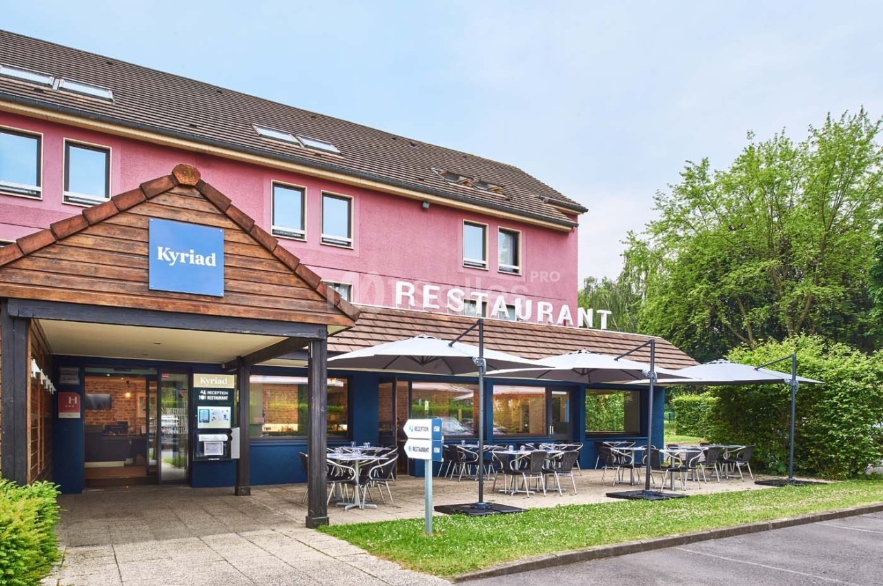 Façade d'un hôtel-restaurant avec terrasse équipée de tables et parasols, entourée de verdure.
