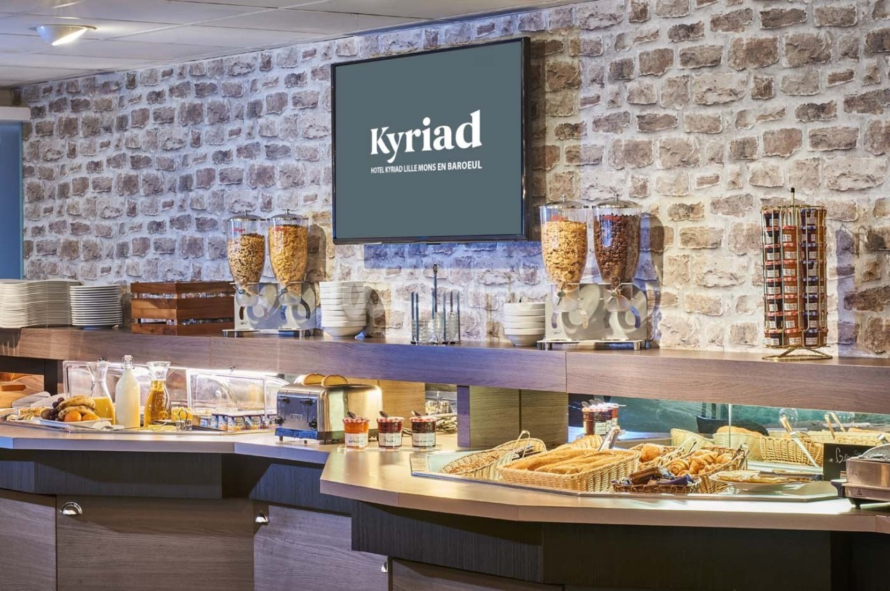 Buffet de petit-déjeuner avec céréales, viennoiseries, pains et boissons, devant un mur en briques.