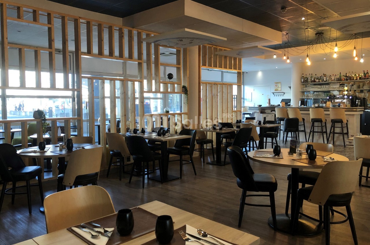 Salle de restaurant moderne avec tables dressées, chaises en bois, bar en arrière-plan et éclairage suspendu.