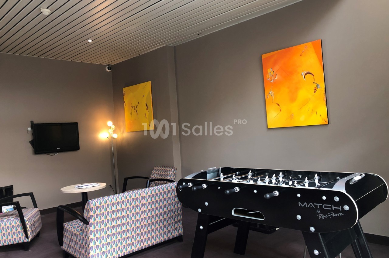 Salle de détente avec baby-foot, fauteuils, table basse, télévision murale et tableaux colorés accrochés au mur.
