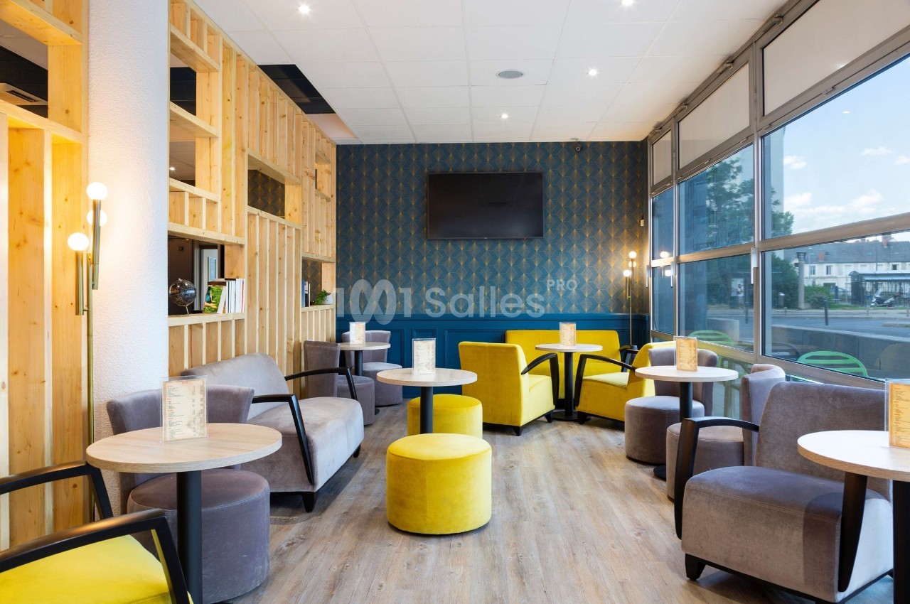 Espace lounge lumineux avec fauteuils gris et jaunes, tables rondes en bois, mur décoratif et grande baie vitrée.