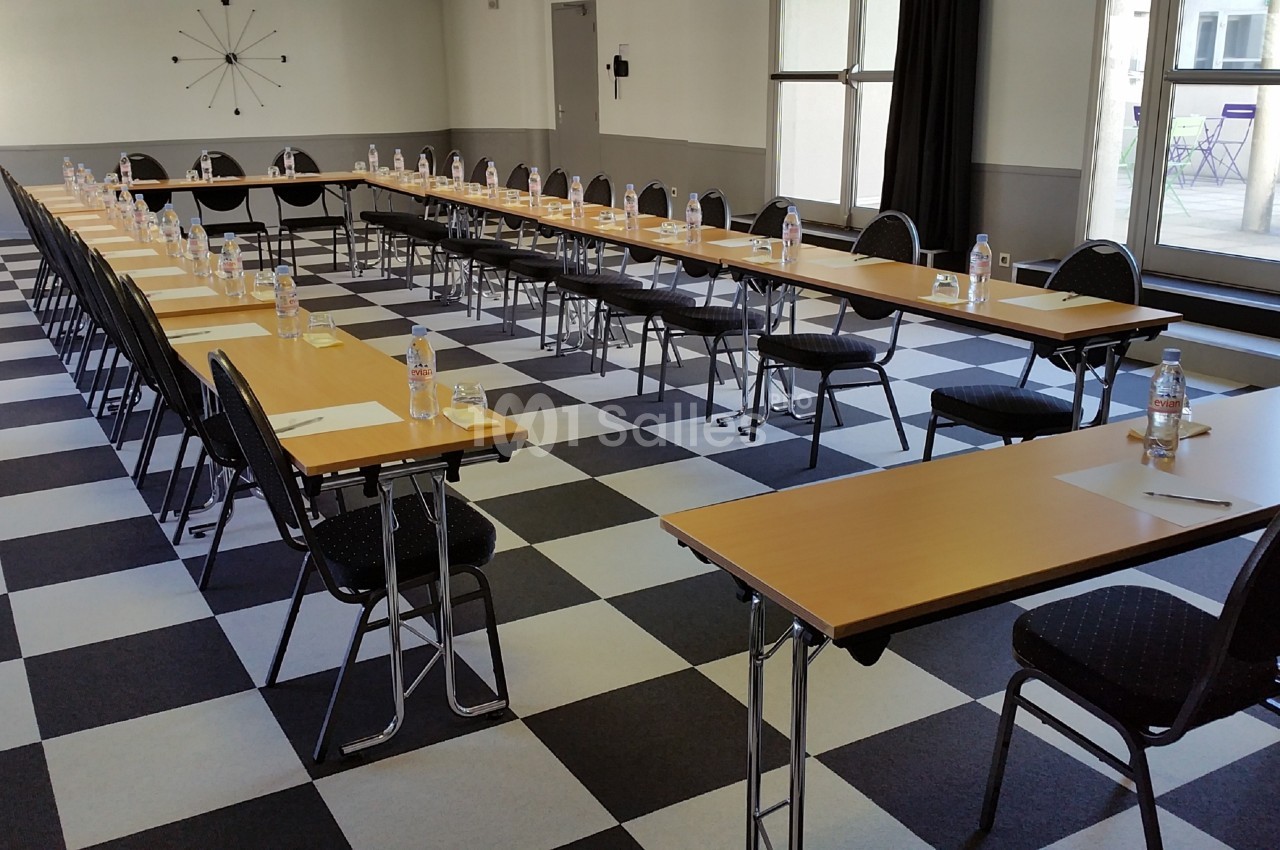 Salle de réunion avec tables disposées en U, chaises noires, bouteilles d'eau et blocs-notes sur un sol à carreaux.