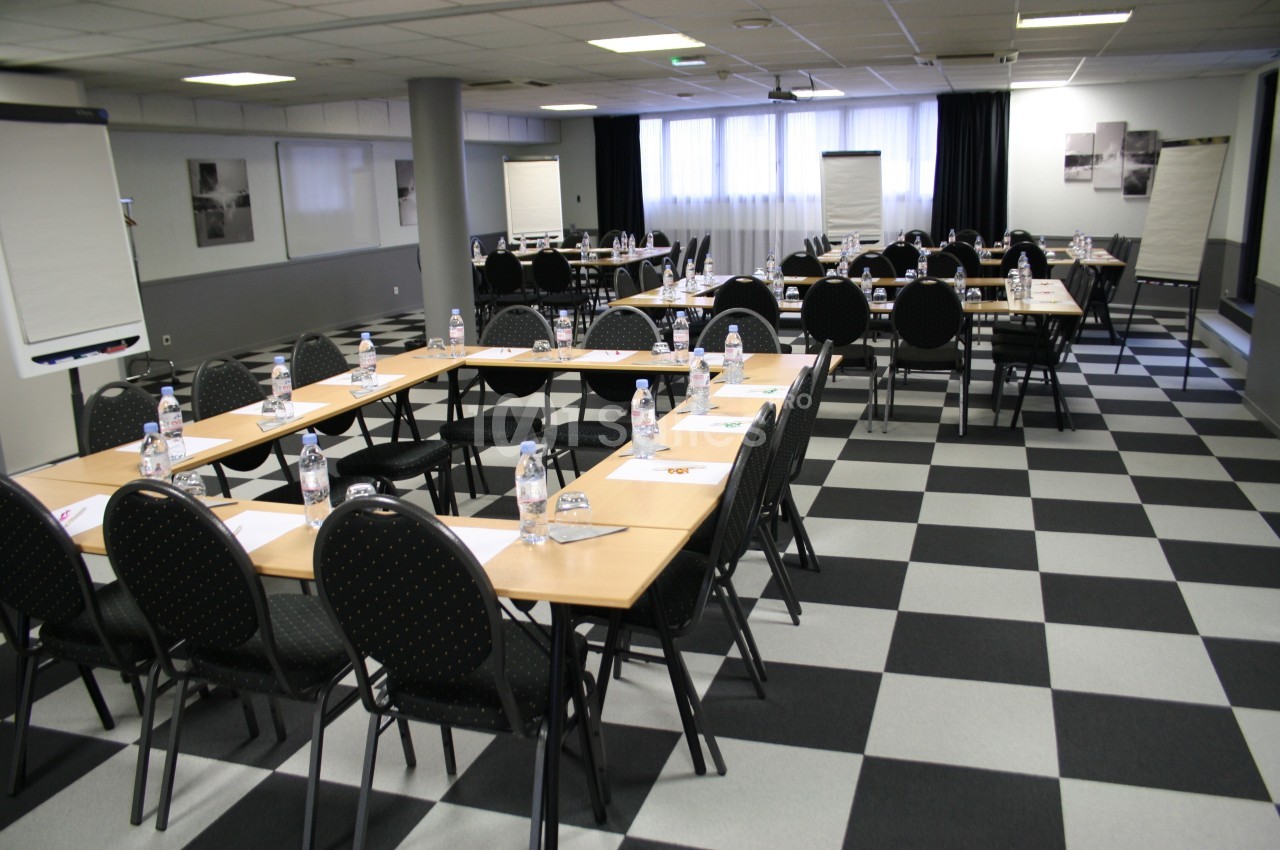 Salle de réunion équipée de tables disposées en U, chaises noires, bouteilles d'eau et chevalets de conférence.