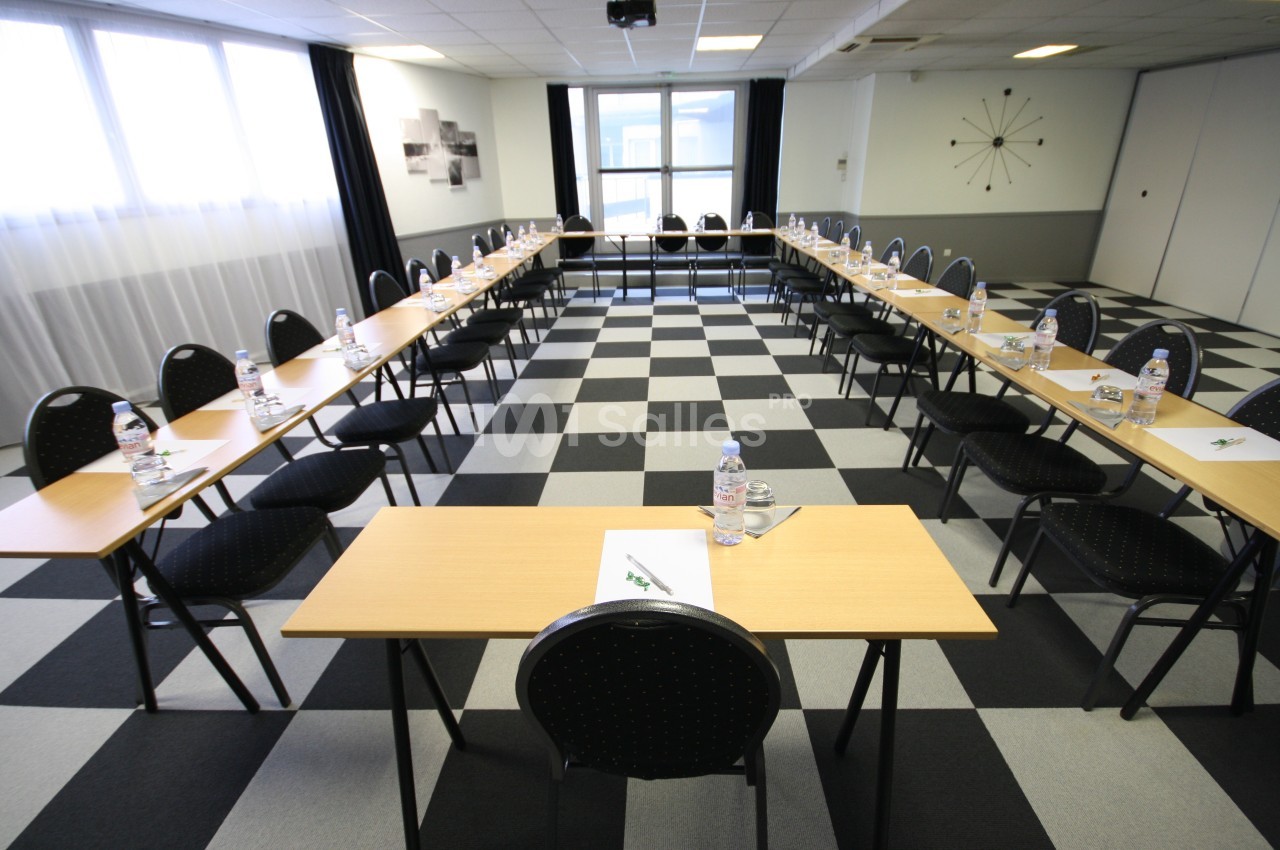 Salle de réunion lumineuse avec tables disposées en U, chaises noires et bouteilles d'eau sur chaque table.