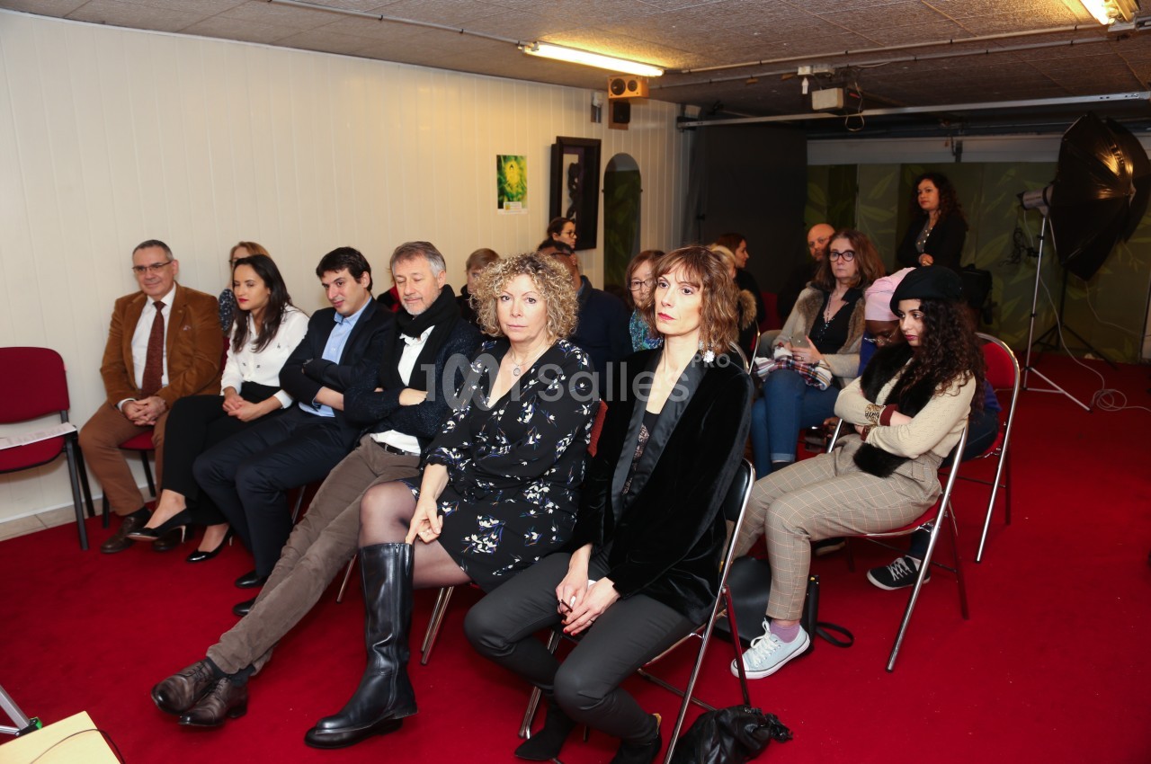 Des personnes assises sur des chaises dans une salle avec moquette rouge, attentives à un événement ou une présentation.