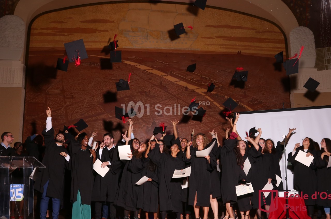 Des étudiants en toges noires célèbrent leur remise de diplômes en lançant leurs mortiers en l'air dans une salle décorée.