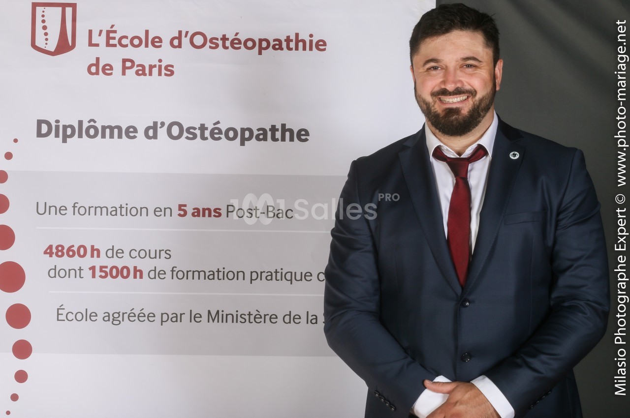Un homme en costume souriant devant une affiche présentant une école d'ostéopathie et ses informations clés.