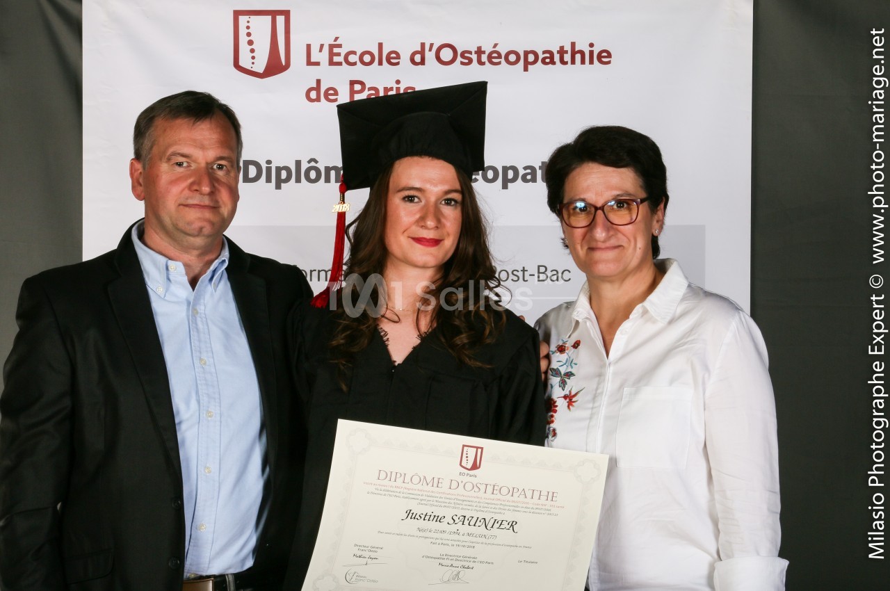 Une diplômée en tenue de cérémonie tient son diplôme, entourée de deux personnes devant une bannière d'école d'ostéopathie.