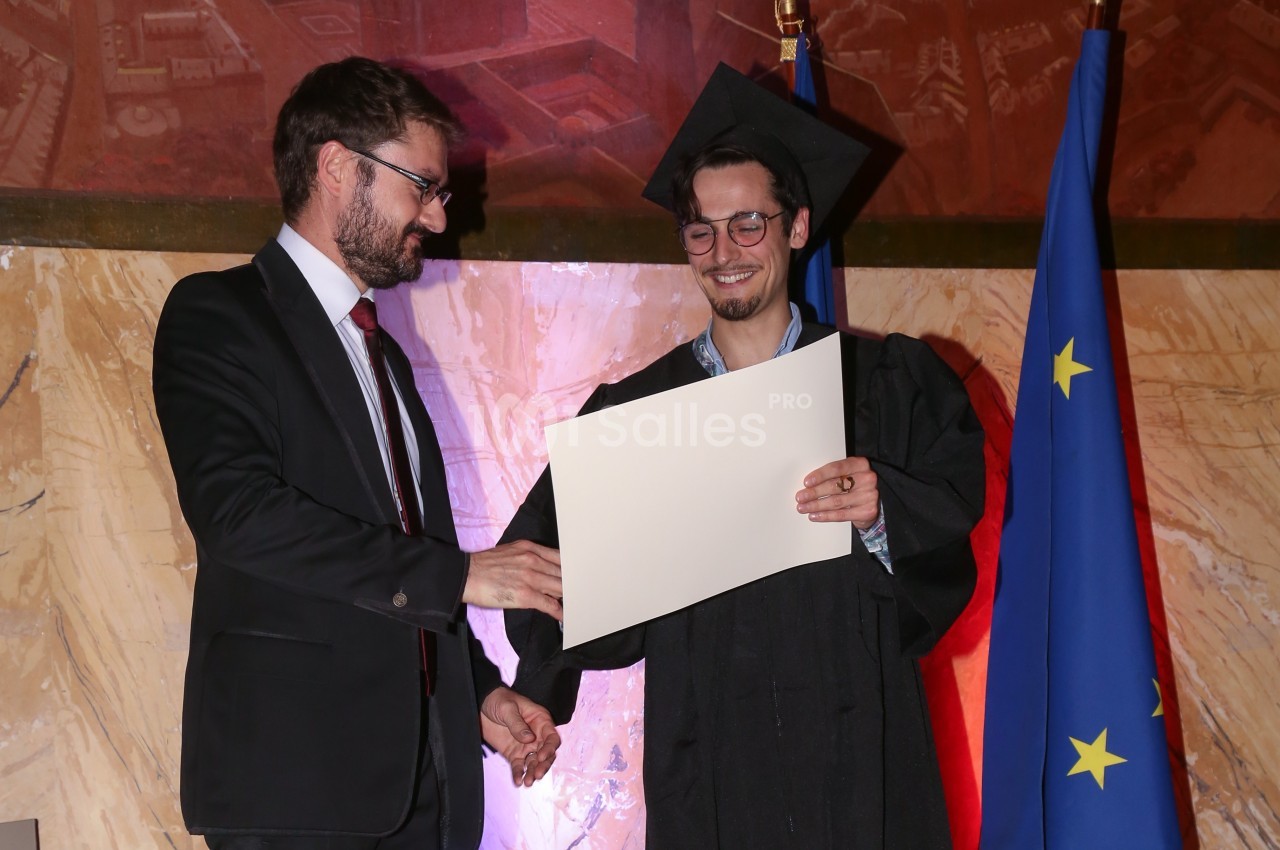 Un diplômé en toge et toque reçoit un certificat des mains d'un homme en costume, devant un drapeau européen.