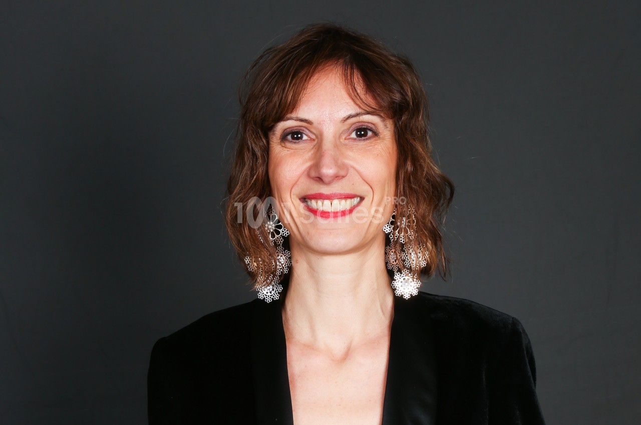 Femme souriante portant des boucles d'oreilles pendantes et une veste noire, sur fond gris uni.