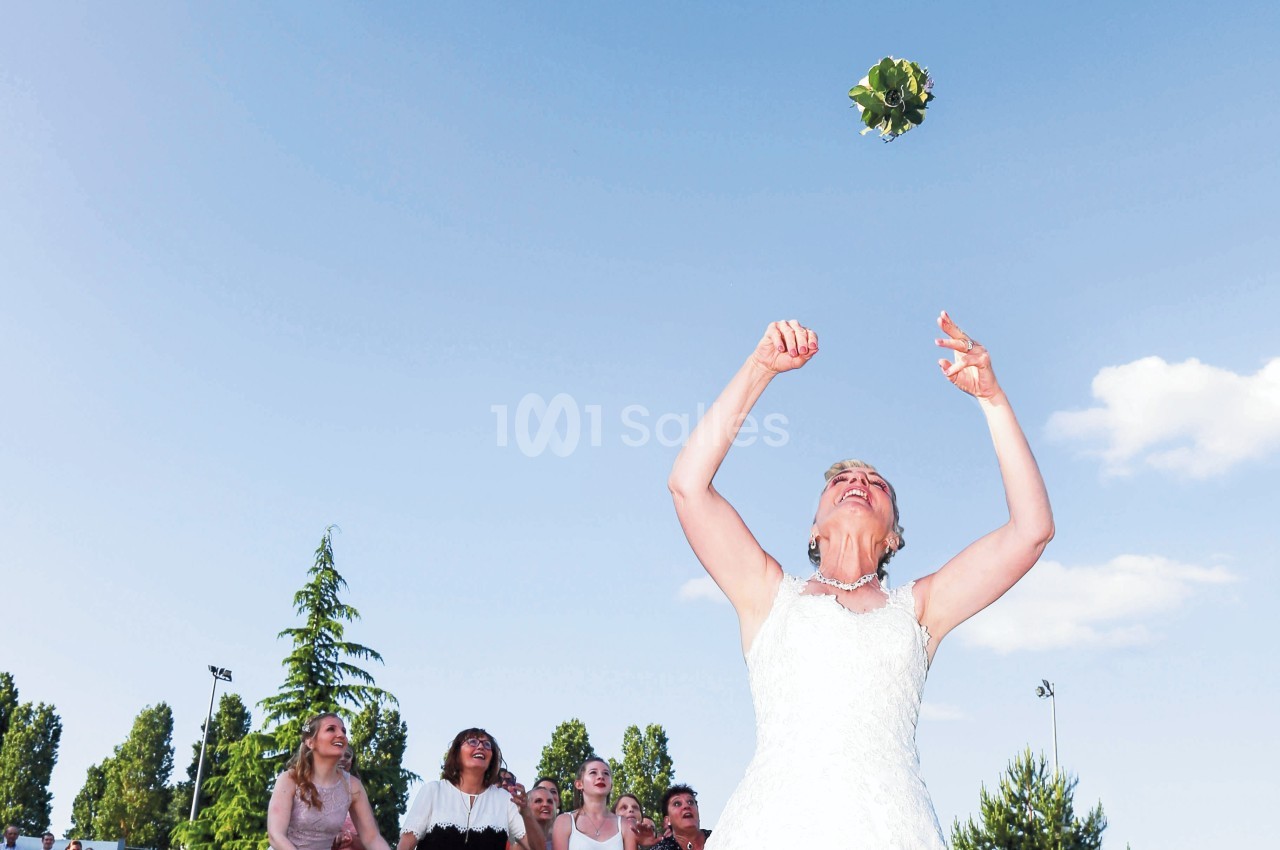 Une mariée lance son bouquet en l'air devant un groupe de femmes en extérieur par temps ensoleillé.