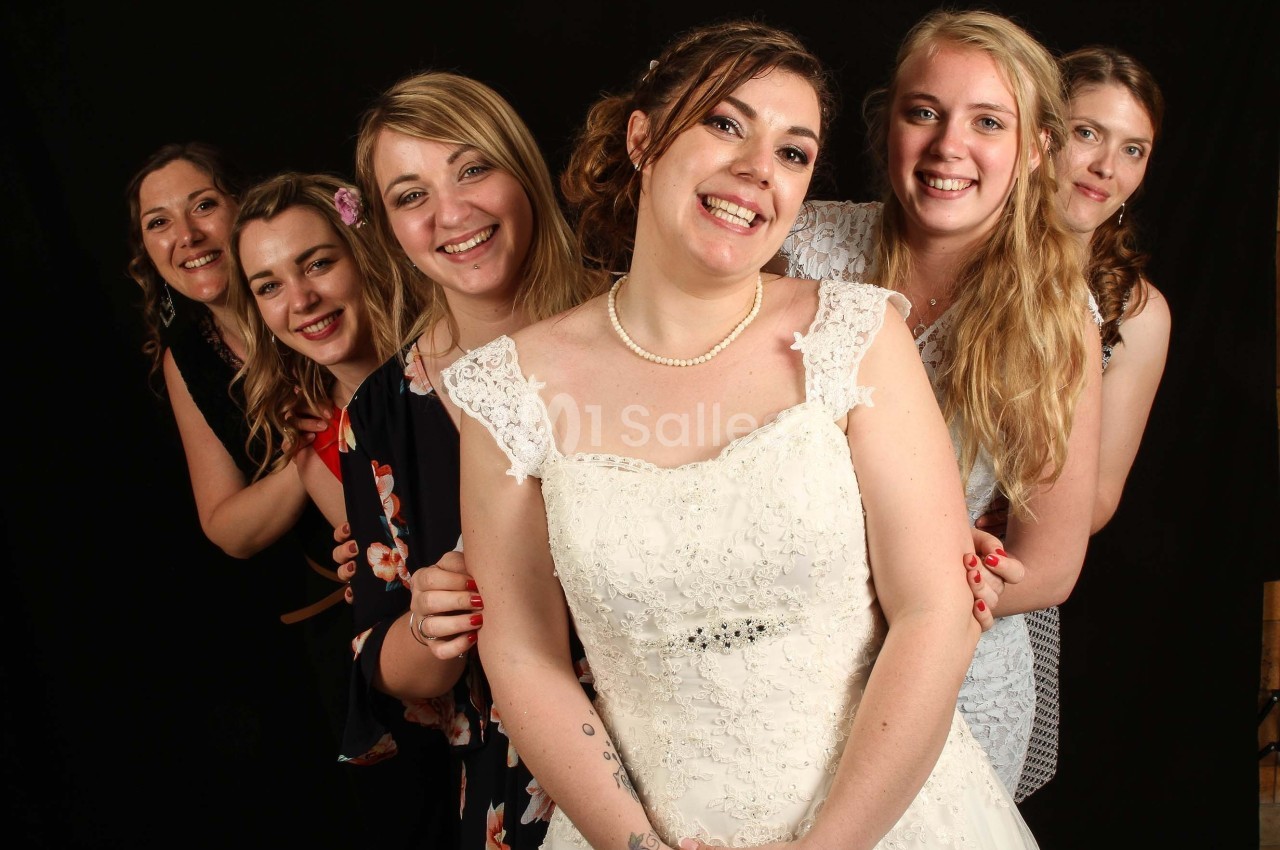 Un groupe de femmes souriantes, dont une en robe de mariée, posant devant un fond noir.
