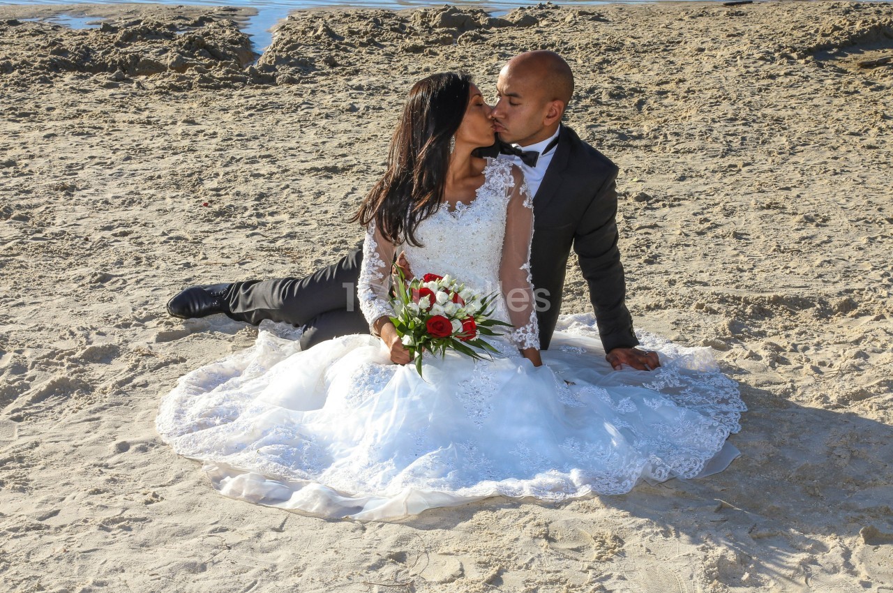 Un couple en tenue de mariage s'embrasse assis sur le sable près de l'eau, la mariée tenant un bouquet de fleurs.