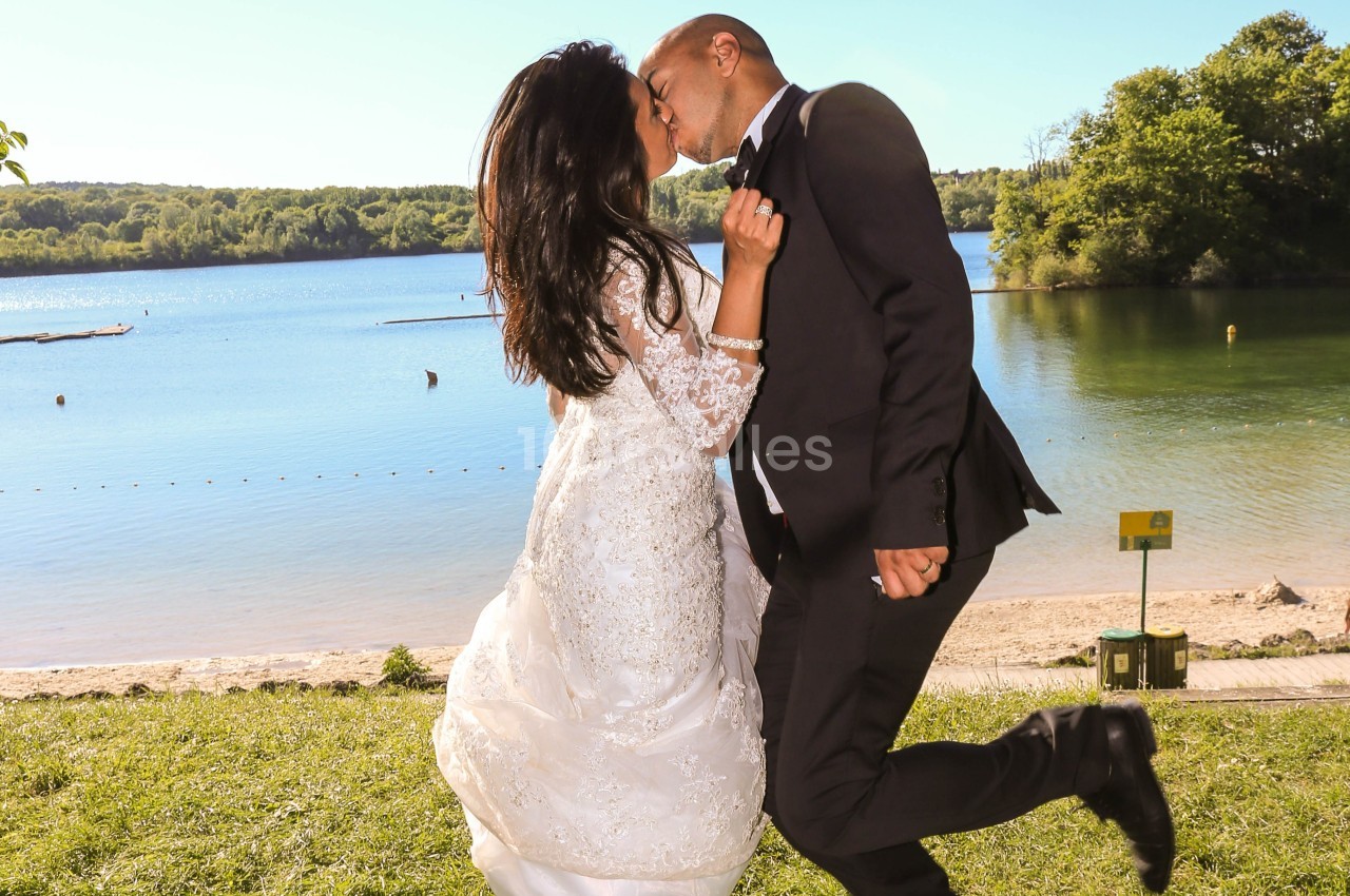 Un couple en tenue de mariage s'embrasse près d'un lac entouré de verdure par une journée ensoleillée.