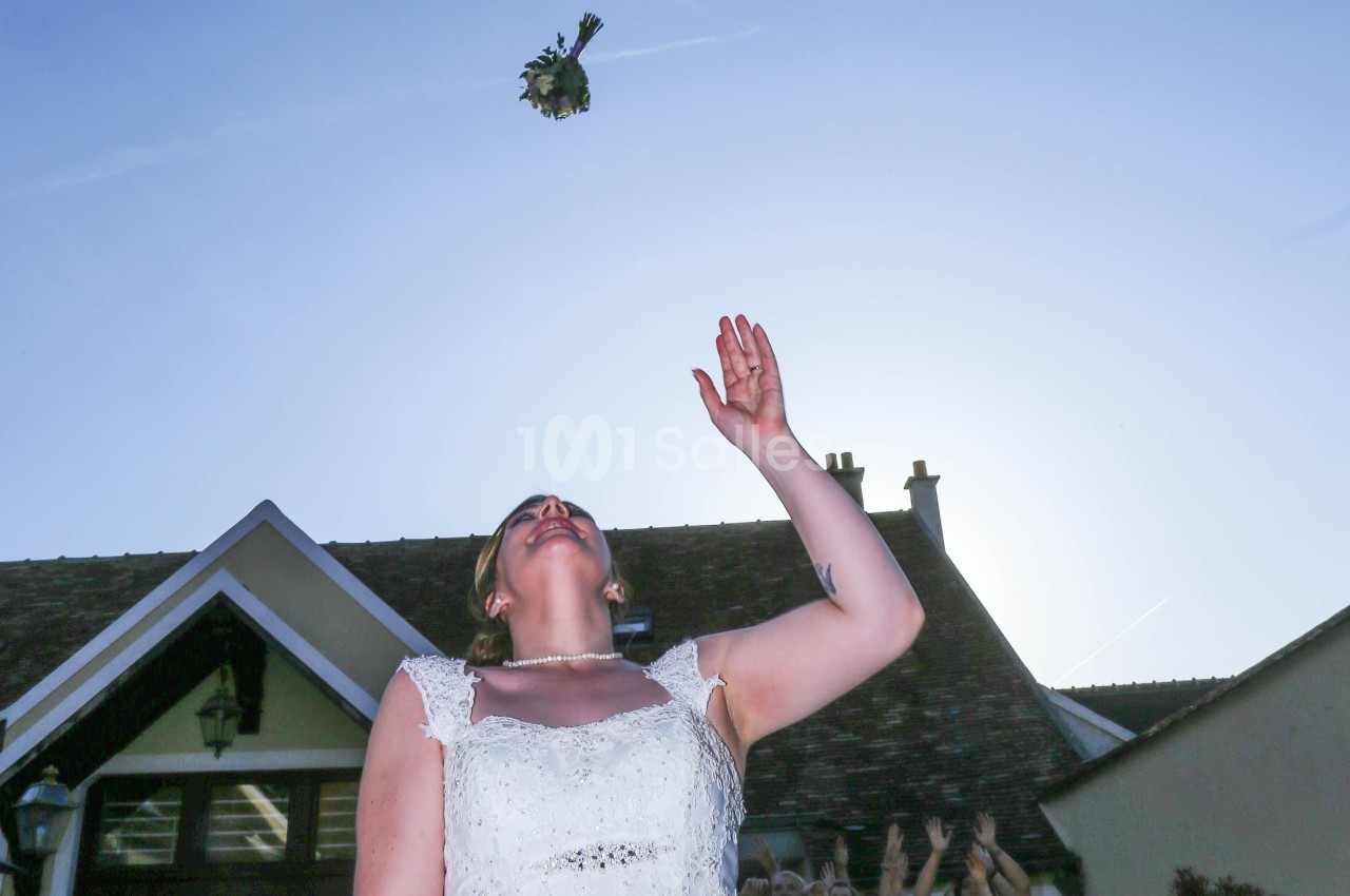 Une mariée lance son bouquet en l'air devant un bâtiment, des mains tendues en arrière-plan.