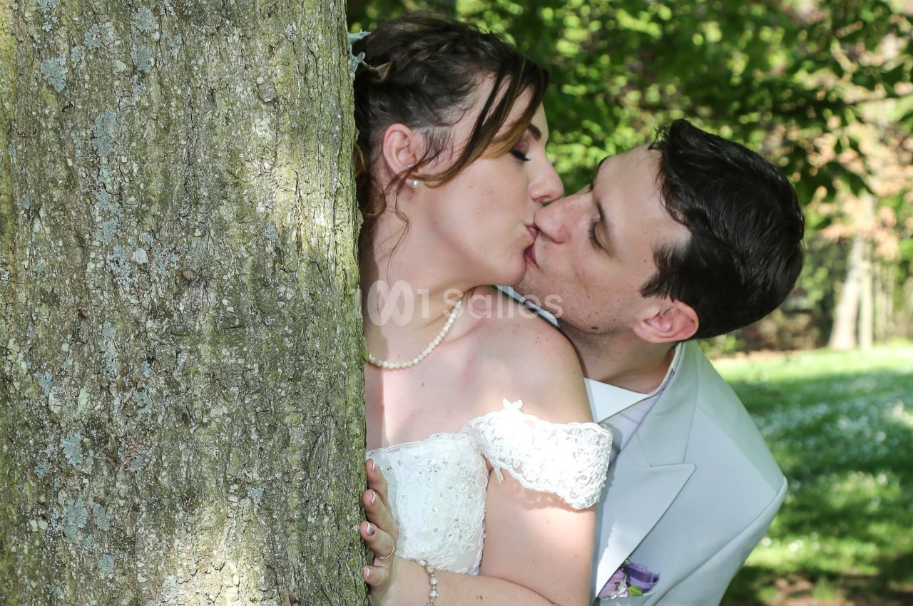Un couple en tenue de mariage s'embrasse tendrement derrière un arbre dans un parc verdoyant.