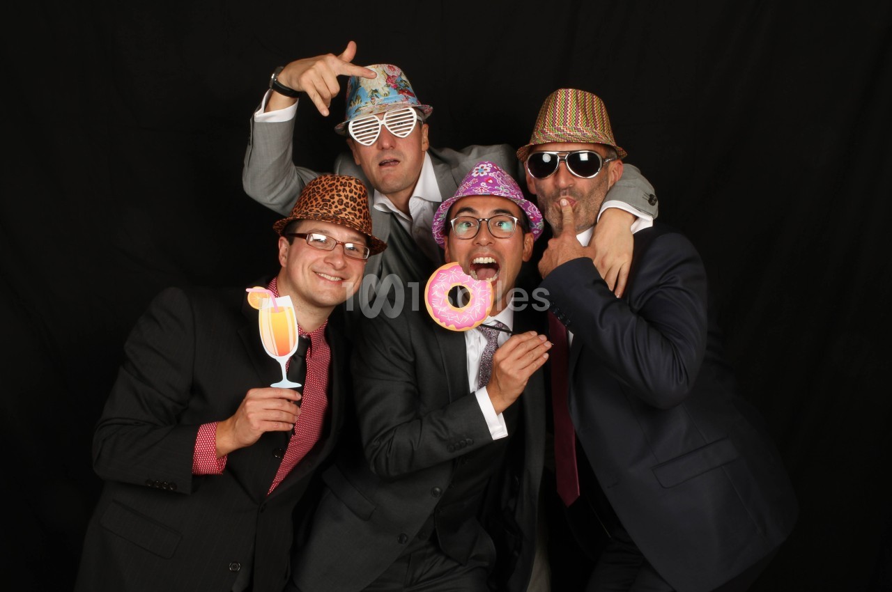 Quatre hommes en costumes posant de manière ludique avec des accessoires festifs sur un fond noir.