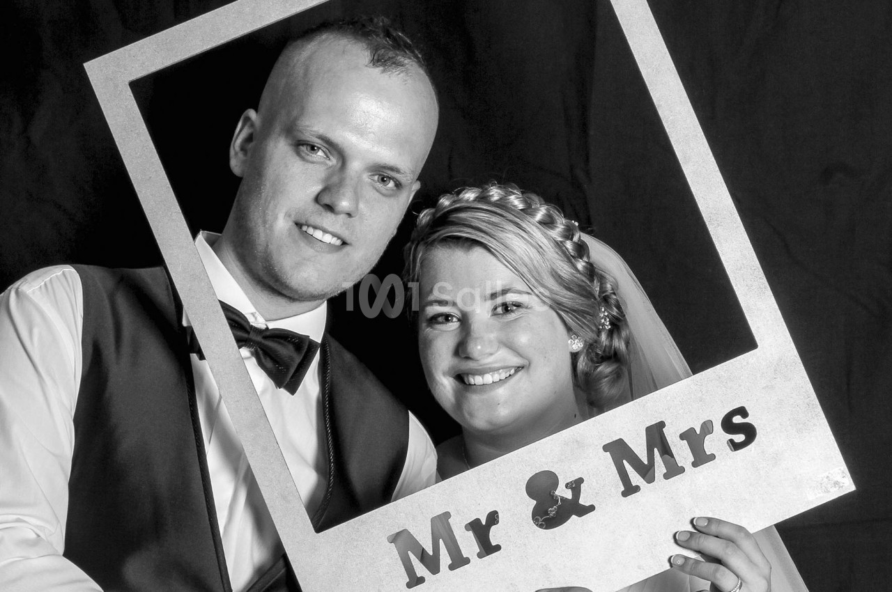 Un couple souriant pose avec un cadre photo portant l'inscription ’Mr & Mrs’ sur un fond sombre.