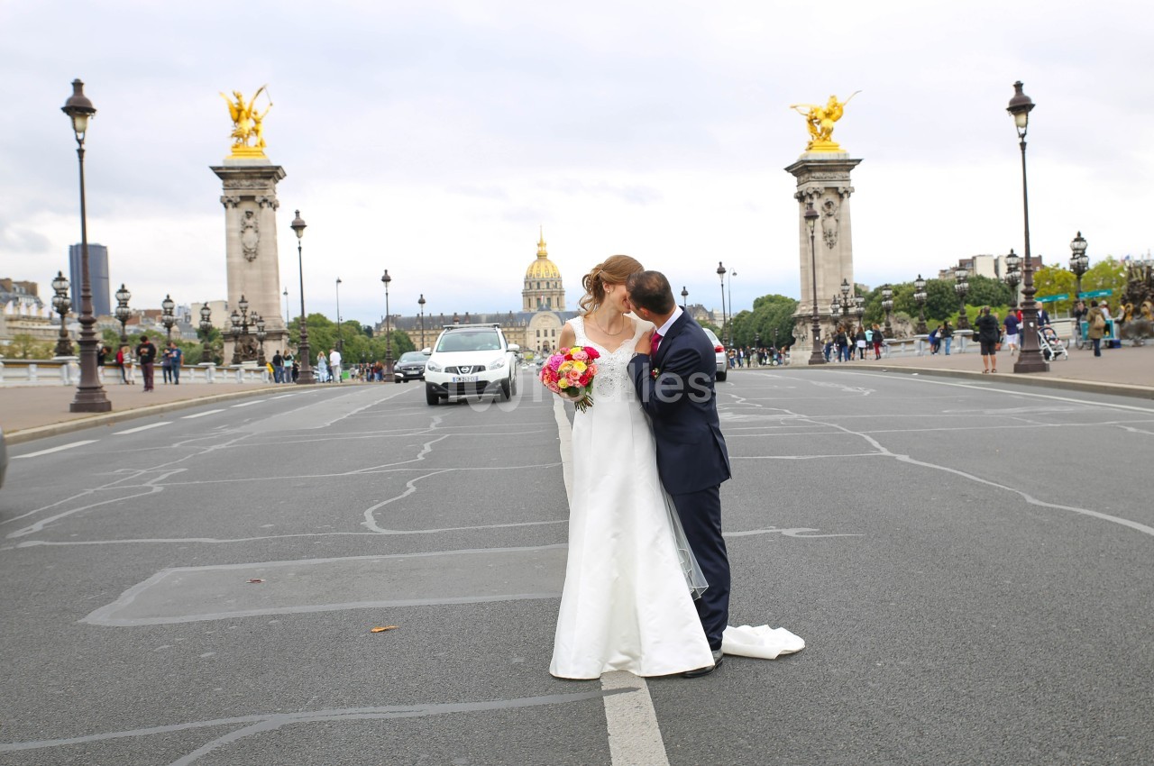 Un couple en tenue de mariage pose sur un pont parisien avec des lampadaires et des statues dorées en arrière-plan.