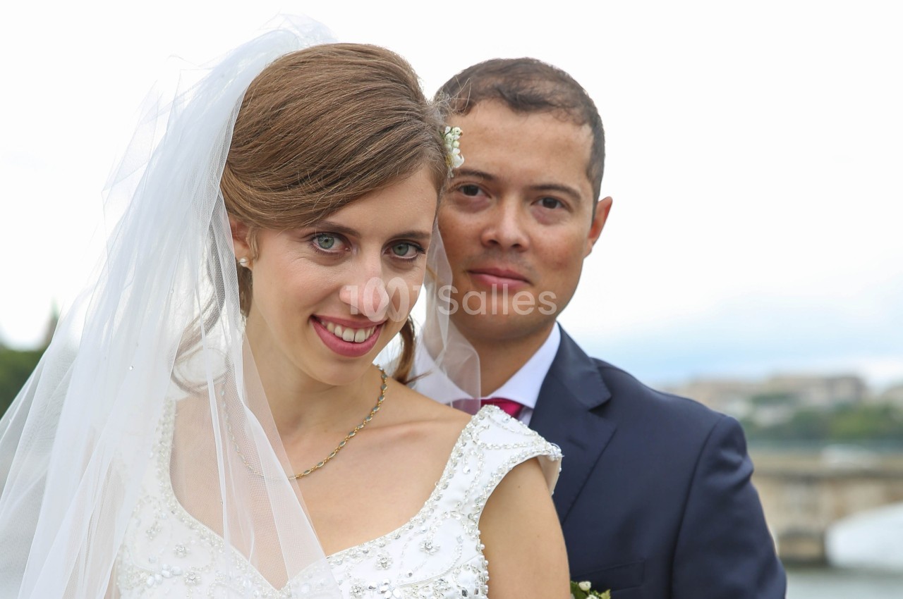 Un couple en tenue de mariage pose en extérieur, la mariée souriante portant un voile blanc.