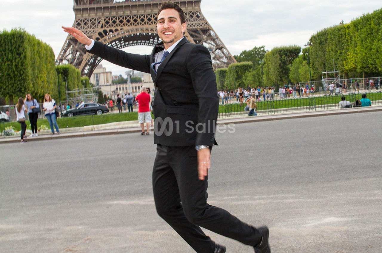 Un homme en costume souriant pose en mouvement devant la tour Eiffel, entouré de visiteurs et d'espaces verts.