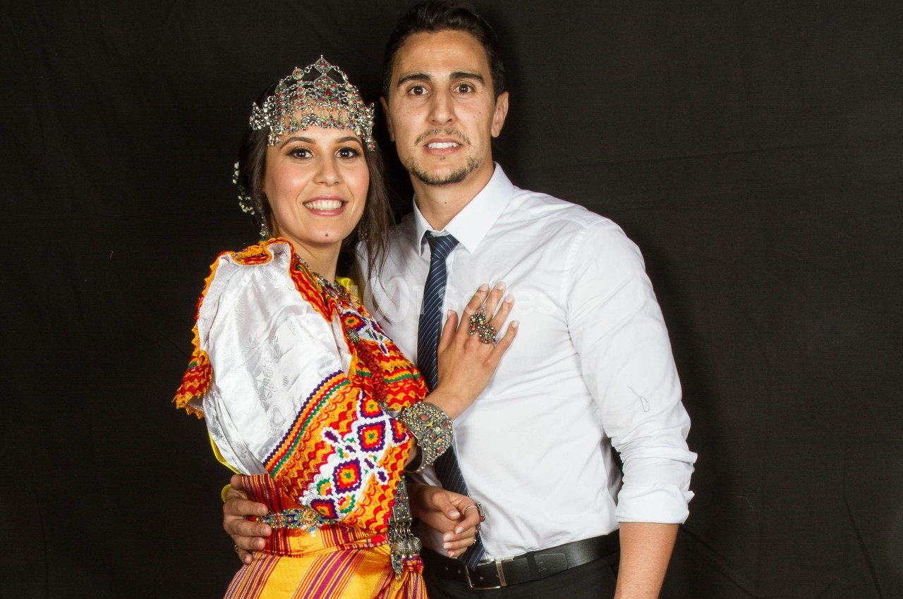 Un couple pose devant un fond noir, la femme porte une tenue traditionnelle colorée et un diadème.