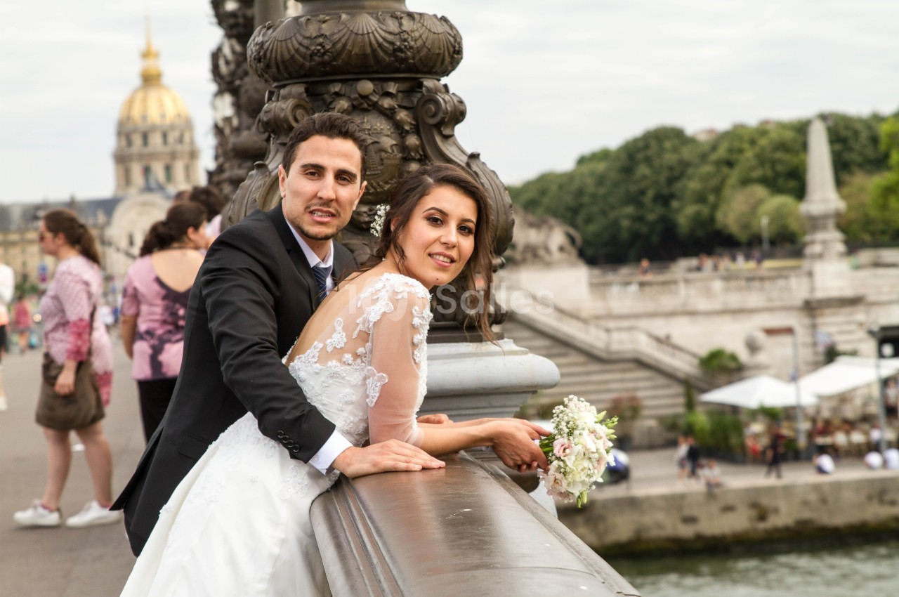 Un couple en tenue de mariage pose sur un pont parisien avec des passants et des monuments en arrière-plan.