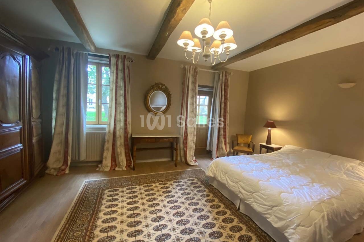 Chambre lumineuse avec lit double, tapis décoratif, armoire en bois, miroir mural et poutres apparentes au plafond.