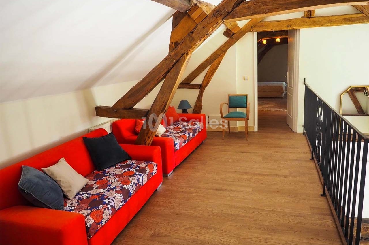 Salon mansardé avec poutres apparentes, deux canapés rouges, parquet en bois clair et chaise bleue près d'une rambarde.