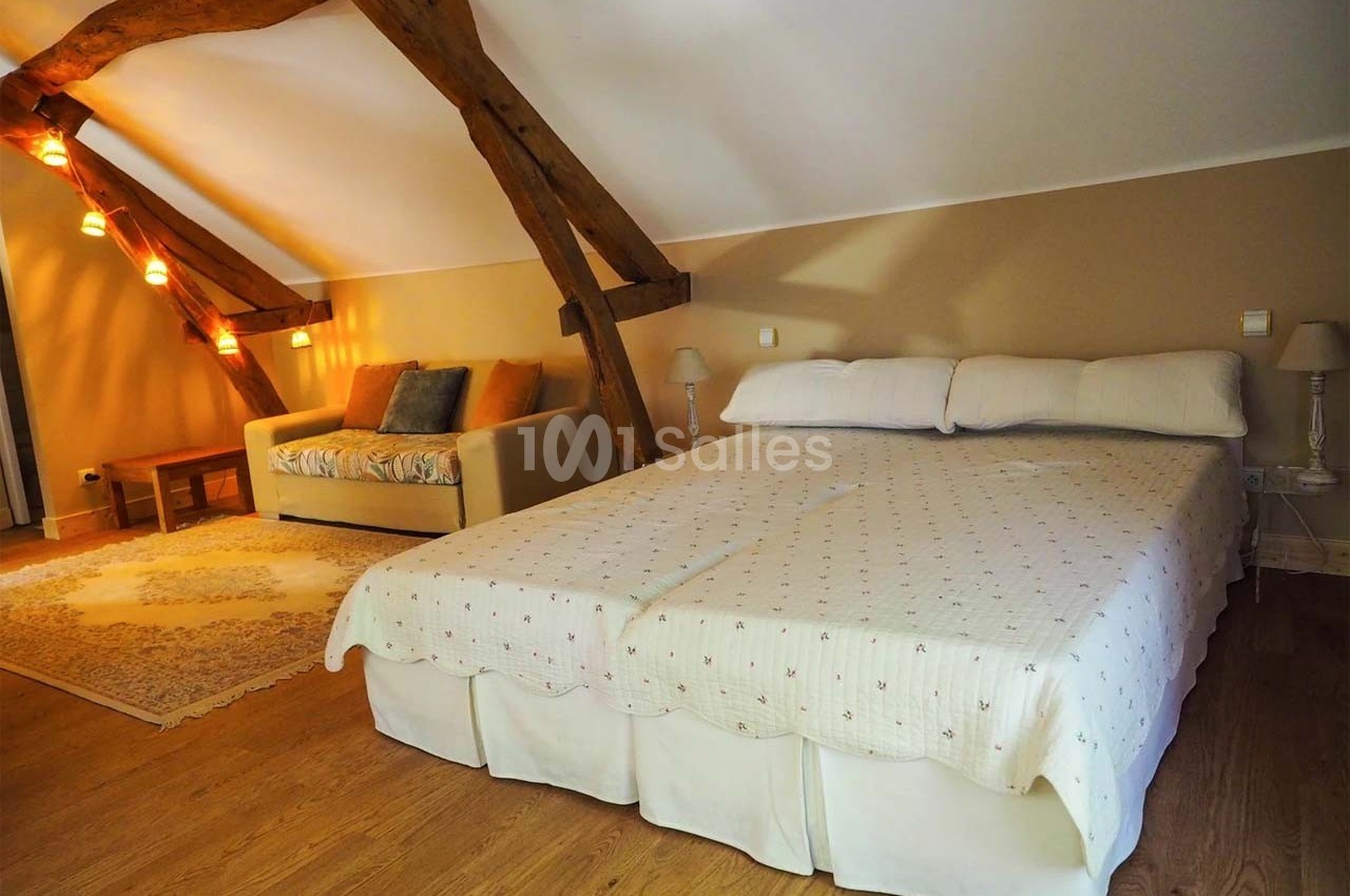 Chambre mansardée avec lit double, canapé, poutres apparentes et éclairage tamisé.