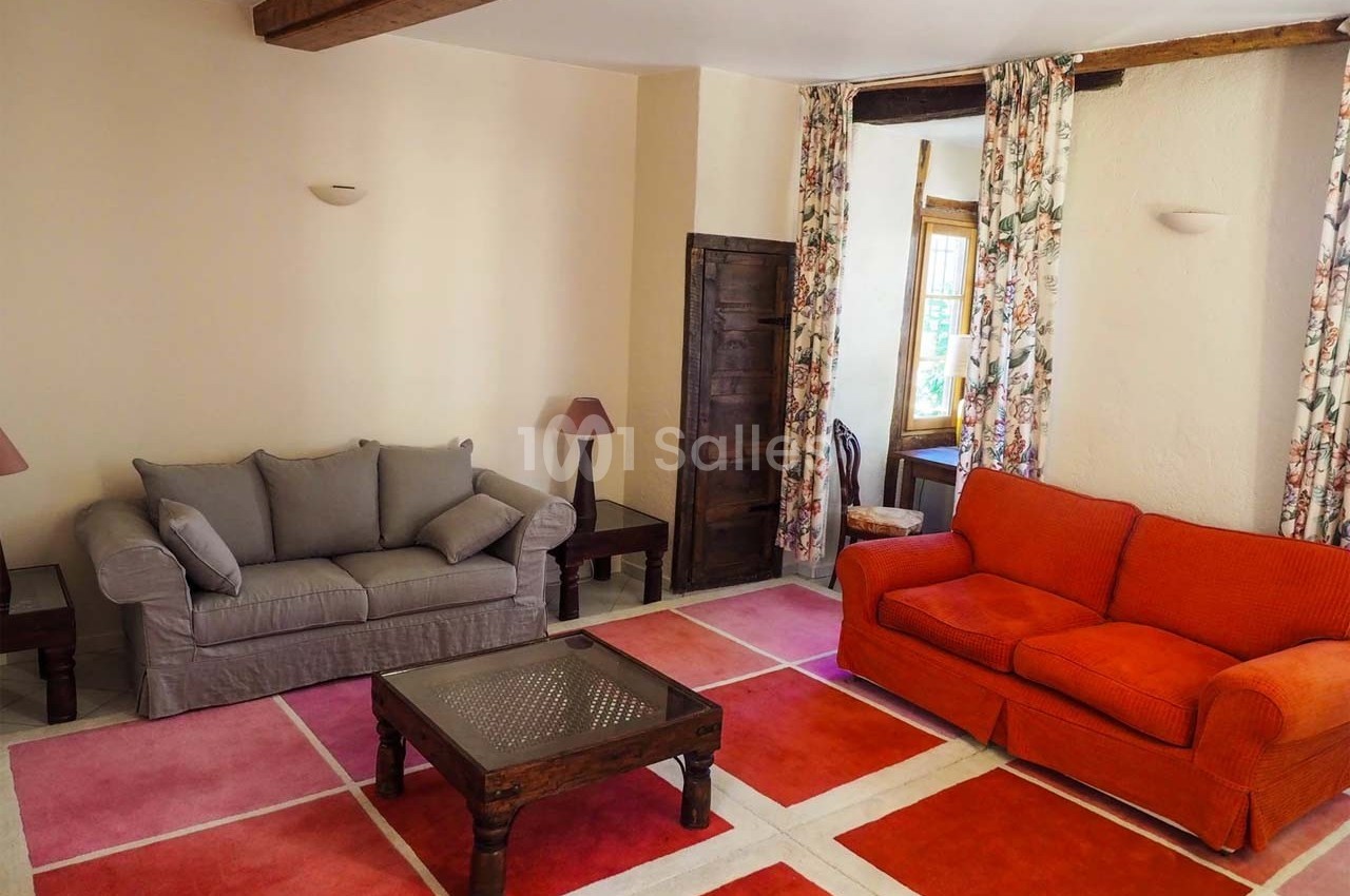 Salon avec deux canapés, table basse en bois, tapis coloré et rideaux fleuris, éclairé par une fenêtre.