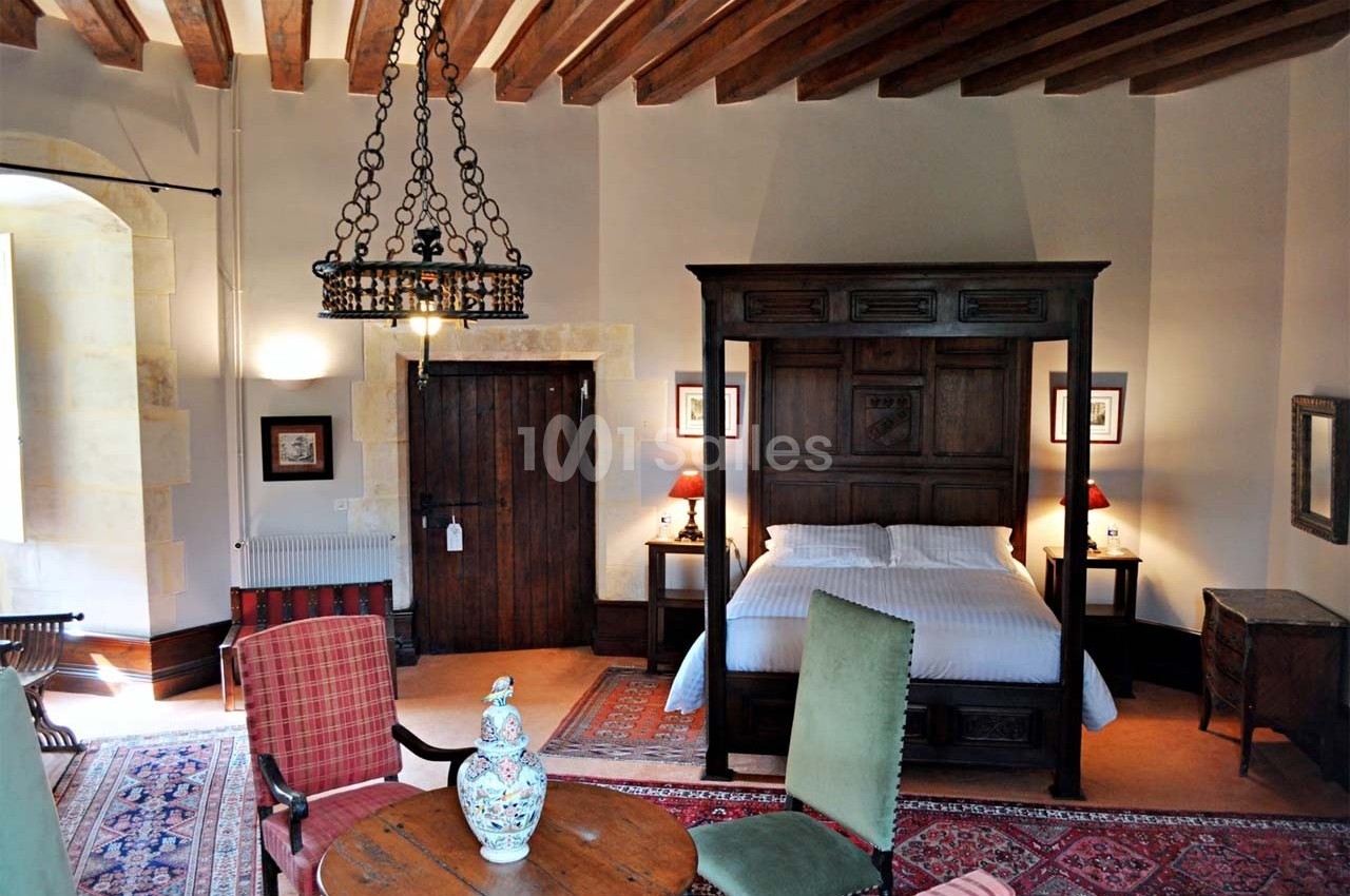 Chambre avec lit à baldaquin en bois, mobilier ancien, tapis colorés et plafond avec poutres apparentes.
