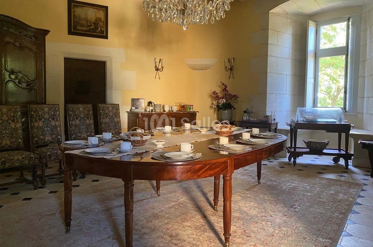Salle à manger lumineuse avec une table dressée pour le petit-déjeuner, entourée de chaises et décorée d'un lustre.