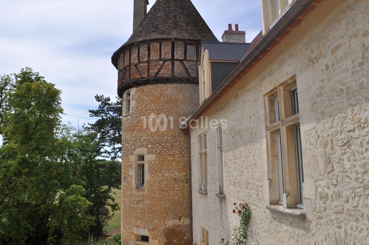 Façade d'un bâtiment ancien en pierre avec une tour ronde, entouré de verdure sous un ciel dégagé.