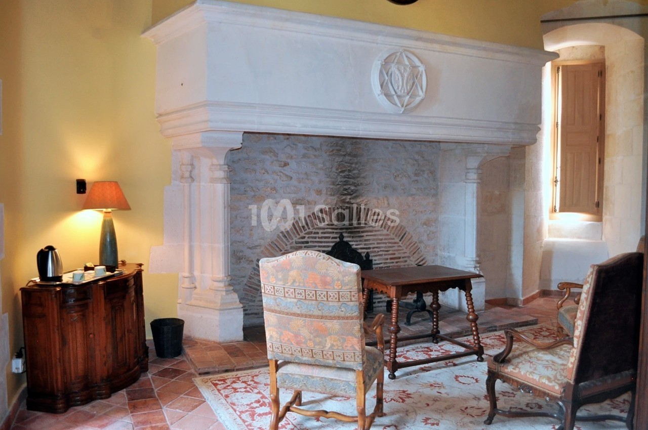 Cheminée en pierre dans une pièce lumineuse avec fauteuils, table basse, lampe et sol en carreaux.
