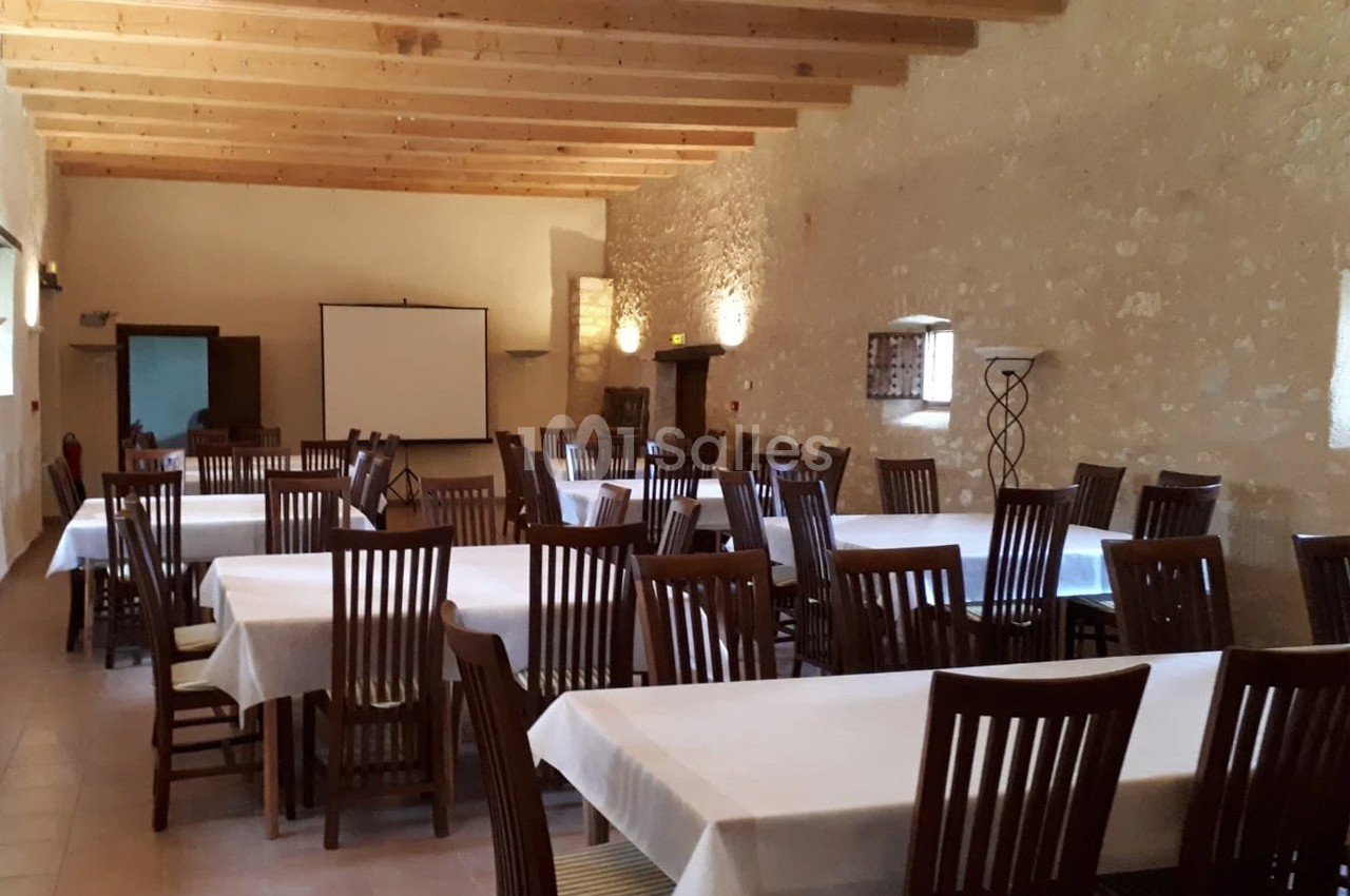 Salle avec des tables rectangulaires couvertes de nappes blanches et des chaises en bois, éclairée par des appliques murales.