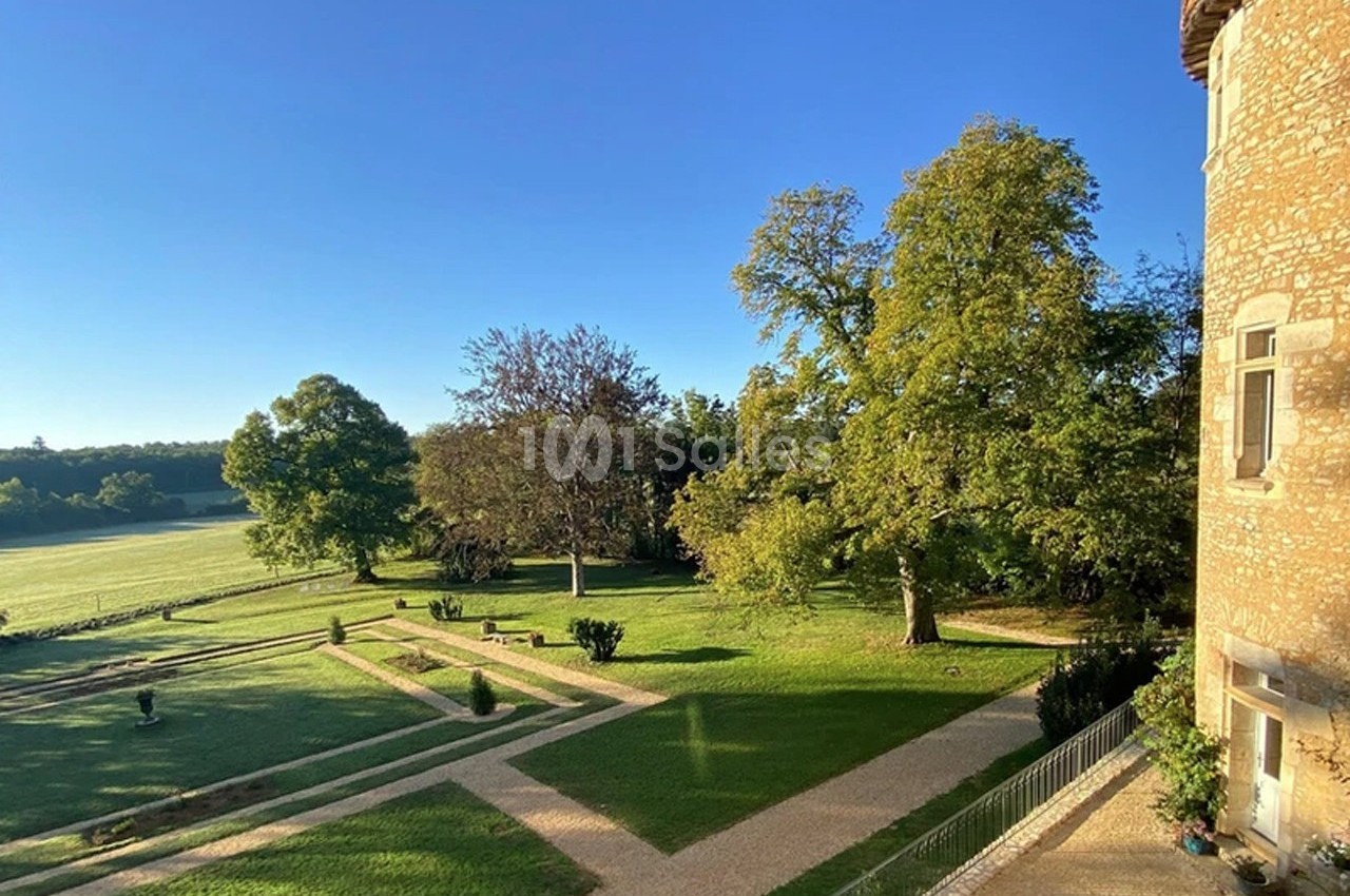 Vue d'un jardin avec allées gravillonnées, pelouse et arbres, bordé par un bâtiment en pierre sous un ciel bleu.