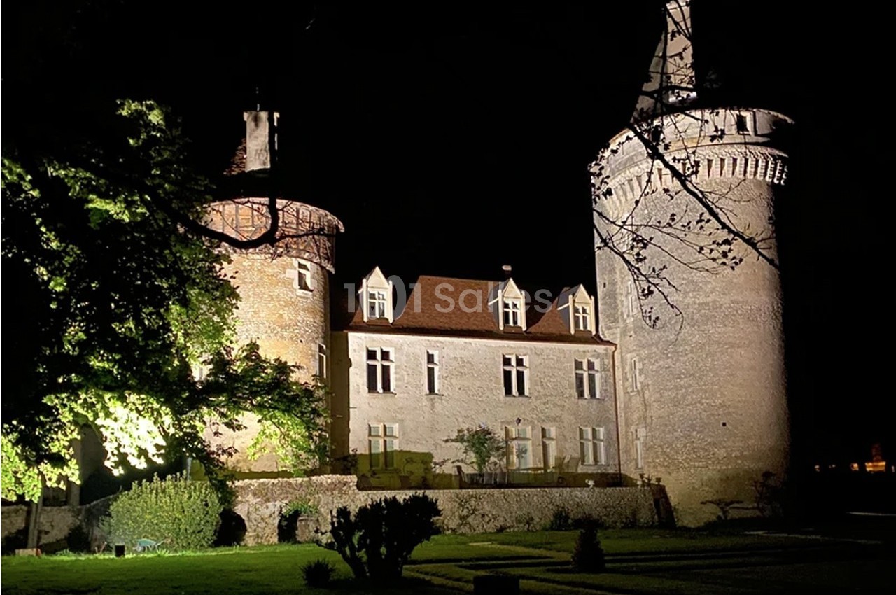 Château médiéval éclairé de nuit, entouré d'arbres et d'un jardin visible au premier plan.