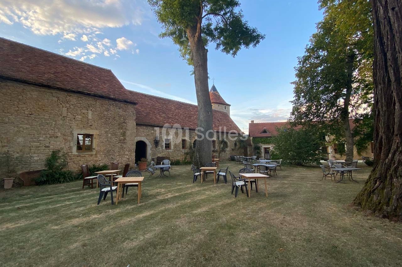 Cour d'une bâtisse en pierre avec tables et chaises disposées sur une pelouse, entourée d'arbres.