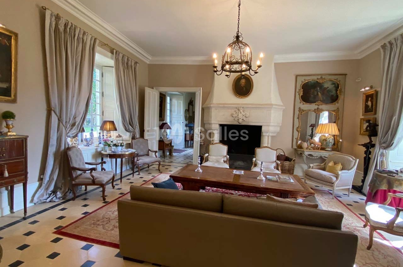 Salon élégant avec cheminée, mobilier classique, lustre en fer forgé, grandes fenêtres et rideaux beiges.
