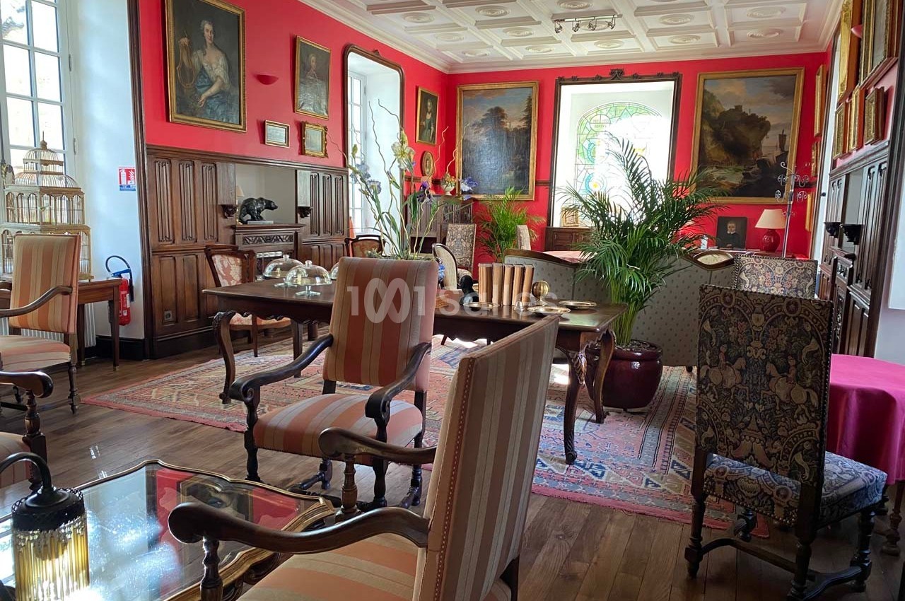 Salon élégant avec mobilier ancien, murs rouges ornés de tableaux, plantes et lumière naturelle entrant par une fenêtre.